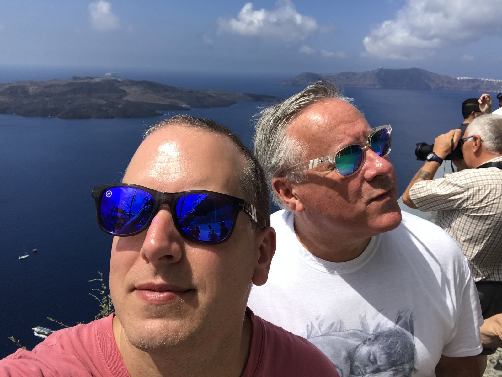JD McArrell and a bandmate overlooking the Santorini caldera, with an unidentified man photographing them from the background.